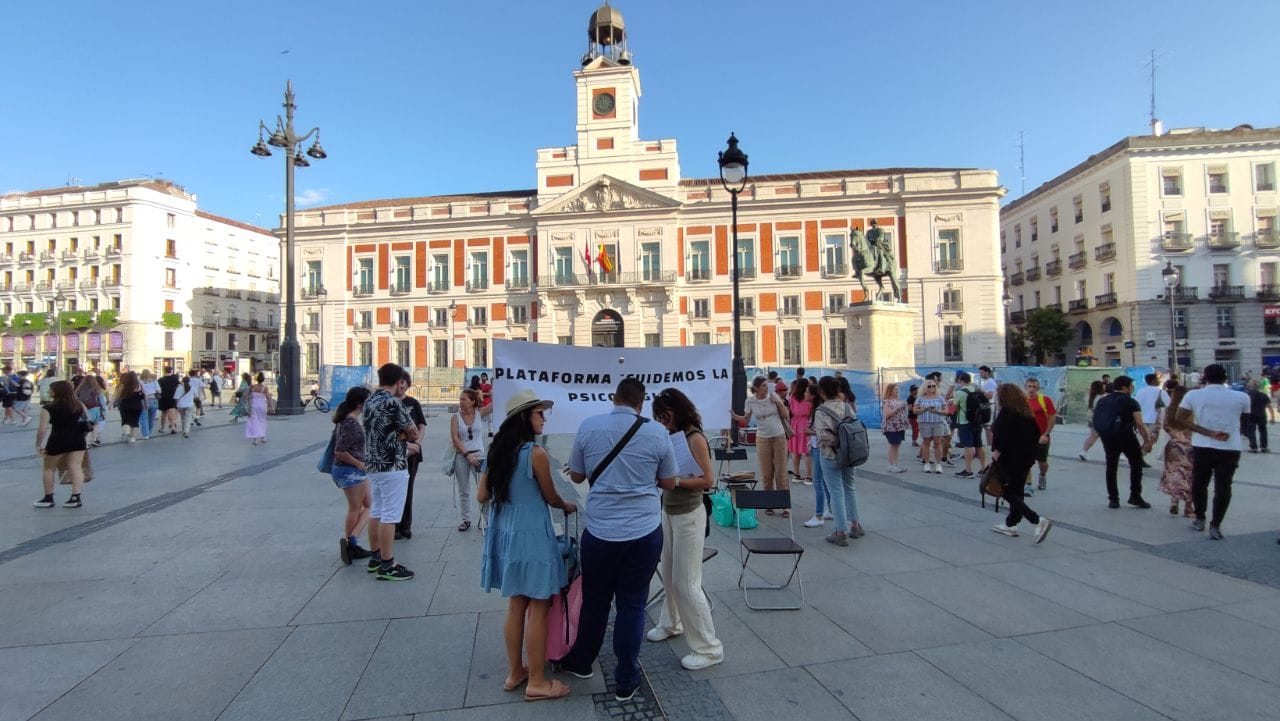 Un miembro de la plataforma habla con varias personas en la Puerta del Sol. De fondo se ve una pancarta desplegada en la que pone "Plataforma Cuidemos la Psicología"